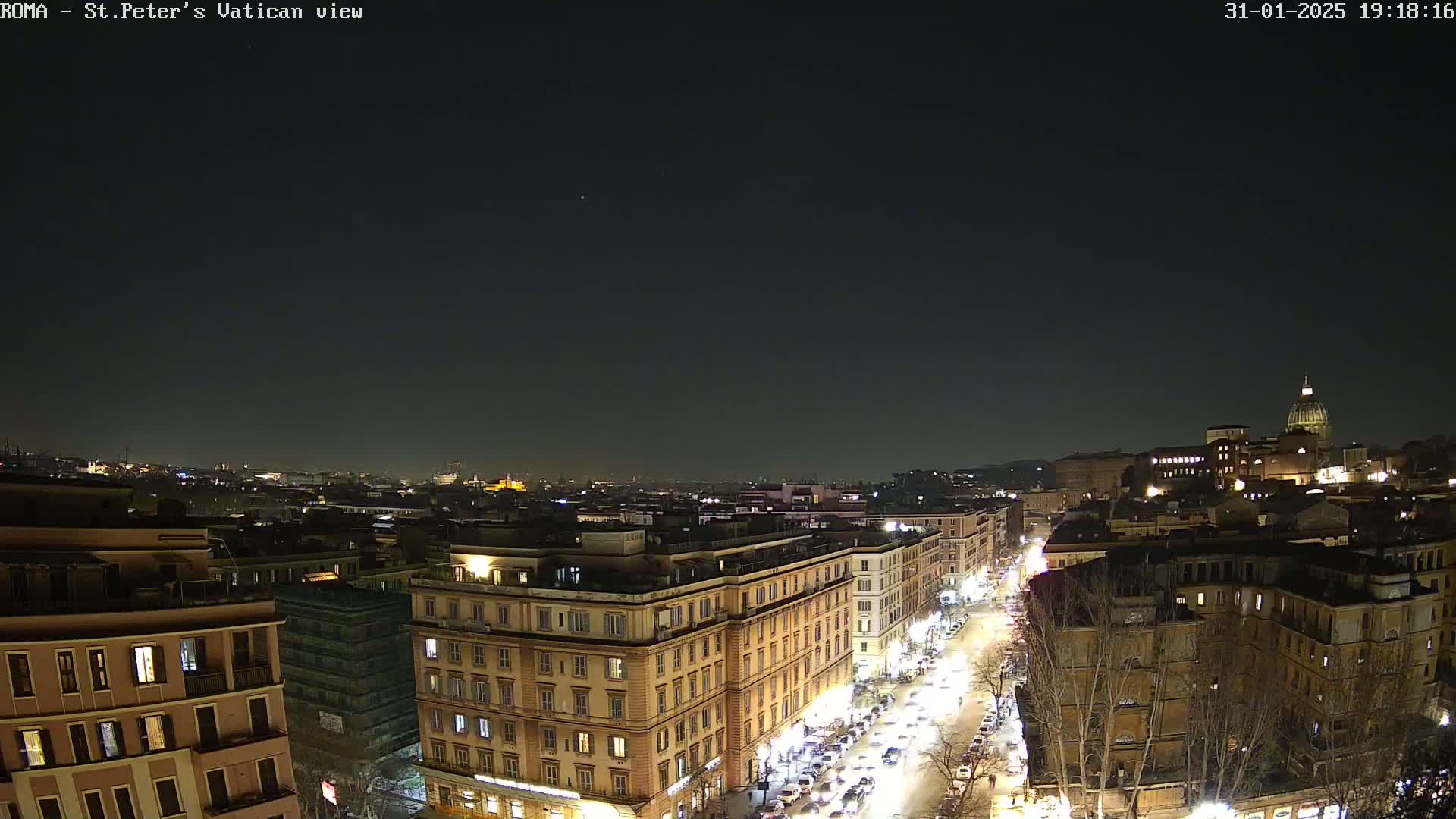 A nighttime, aerial view of a city with illuminated buildings and streets, showing a dome in the distance under a dark sky.