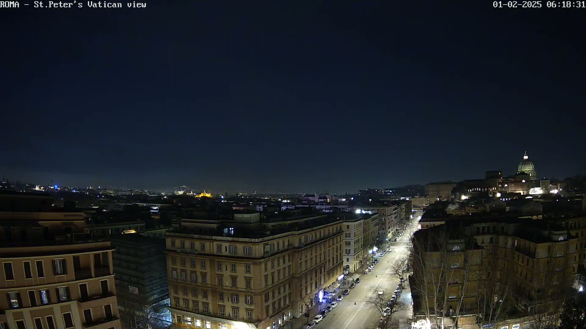 A nighttime, aerial view of a city street lined with buildings, extending toward a larger structure topped with a dome under a clear, dark sky.