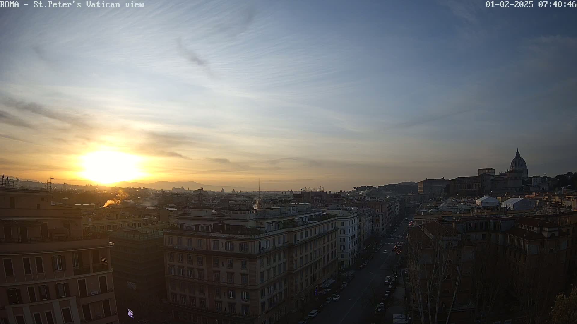 A cityscape at sunrise, with the sun rising behind a distant mountain range, shows buildings and streets below, and St. Peter's Basilica in the distance under a mostly clear sky.