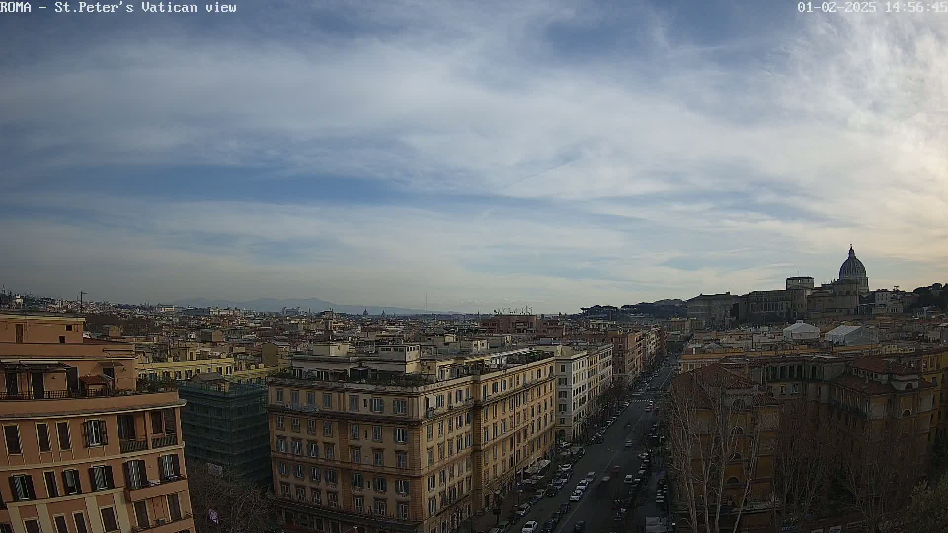 A high-angle, long shot of a cityscape under a mostly cloudy sky shows a street lined with buildings leading toward the Vatican, with distant mountains visible on the horizon.