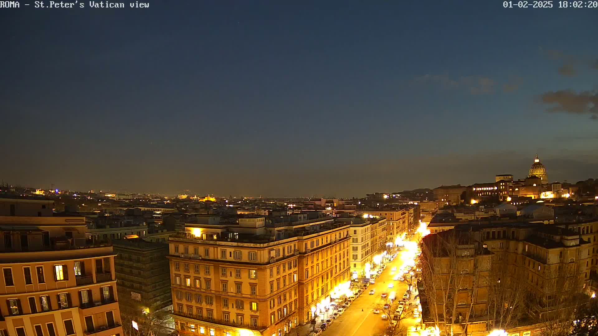 A nighttime view of a city, possibly Rome, showing illuminated buildings and streets leading towards a large domed structure in the distance under a clear, dark sky.