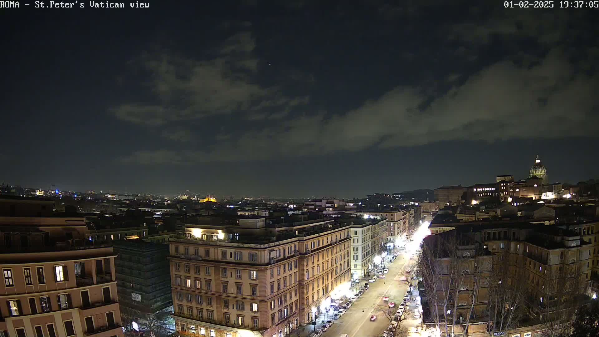 A nighttime, partly cloudy view of a city street scene with buildings illuminated, showing a cityscape extending into the distance and the dome of a large building visible in the upper right.