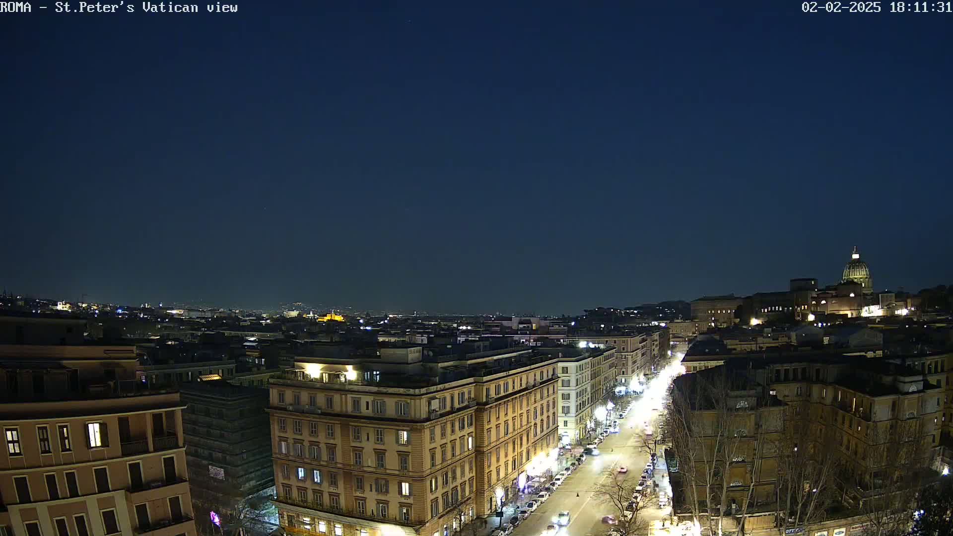 A nighttime, high-angle view of a city street lined with buildings, leading to a large dome-topped structure under a clear, dark sky.