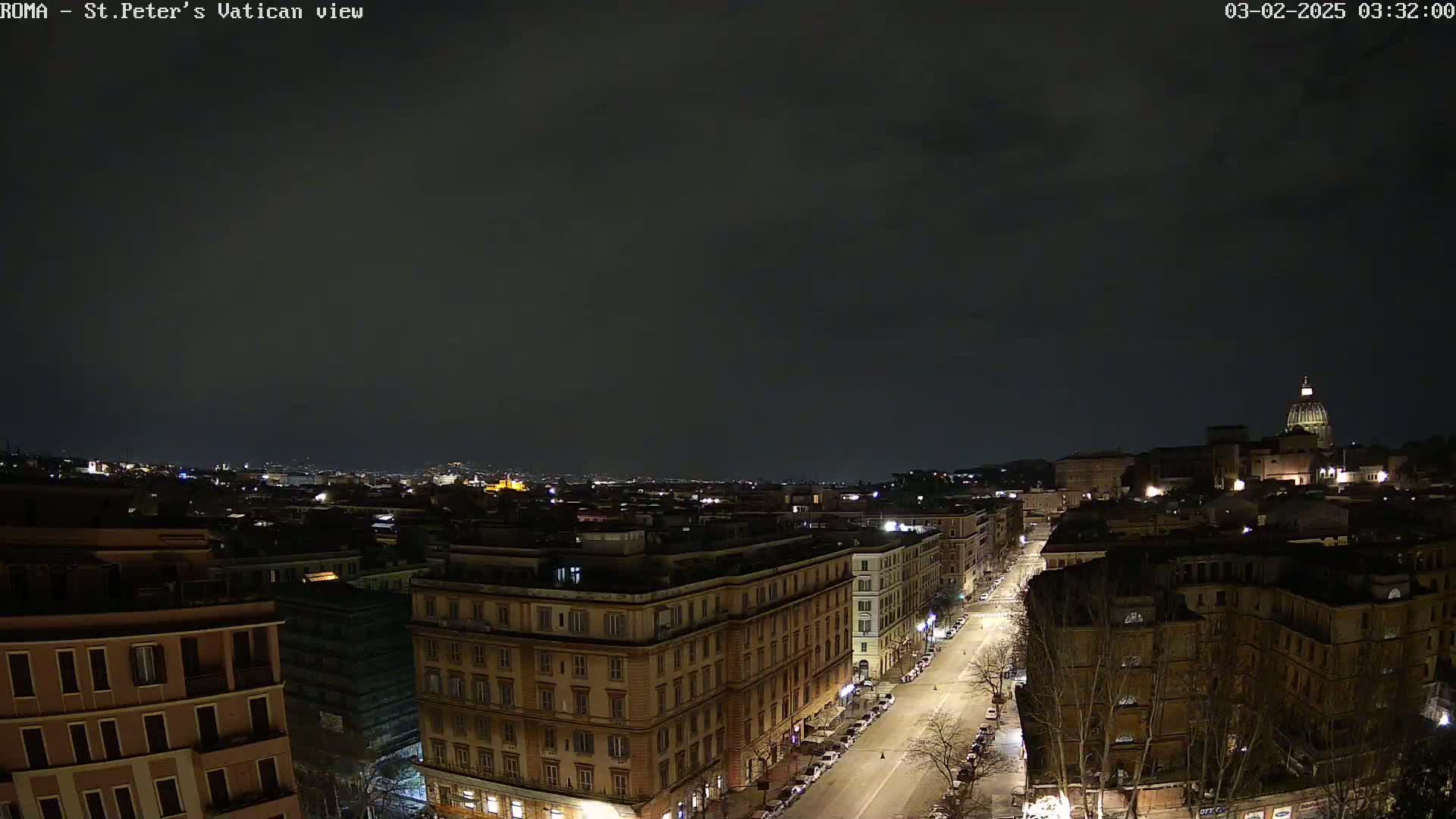 A nighttime view of a city street scene with buildings and a dome-shaped structure visible in the distance under a dark, cloudy sky.