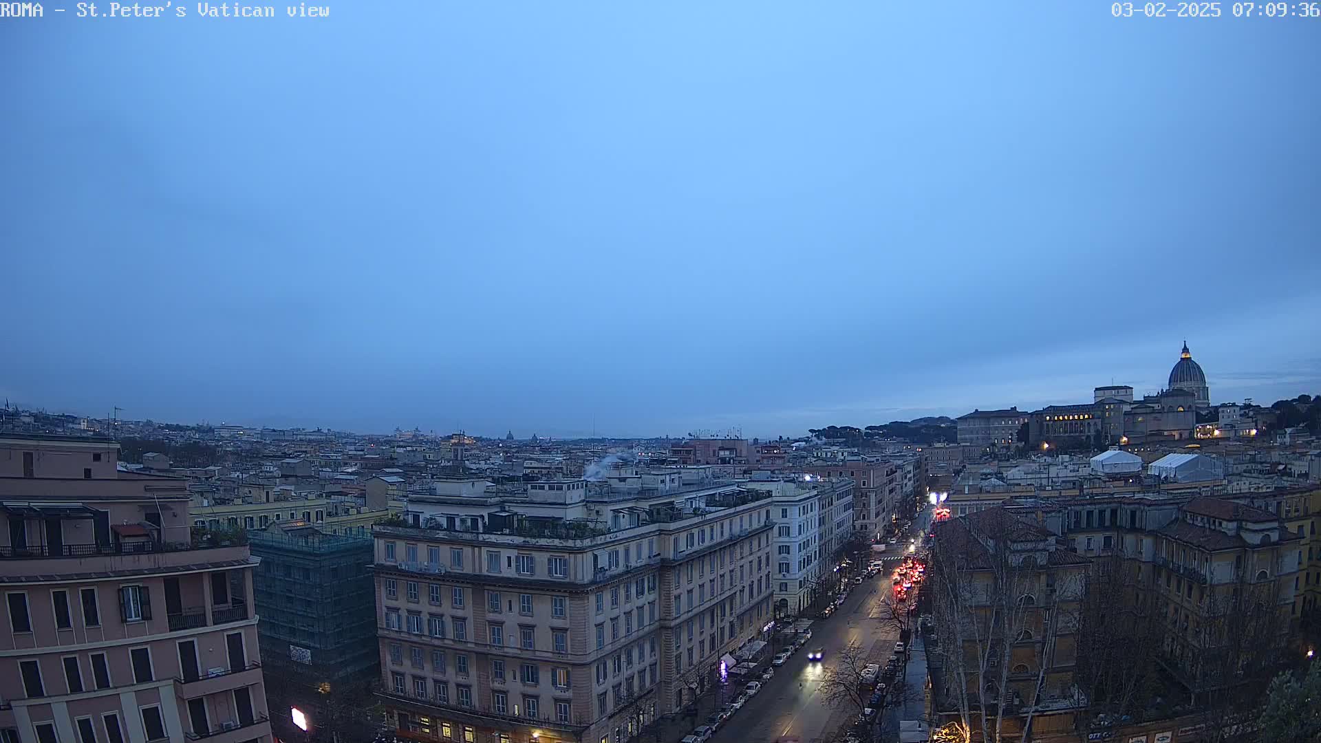 A twilight view of Rome, showing a cityscape with buildings and a street, under a clear, dusky blue sky, with St. Peter's Basilica visible in the distance.