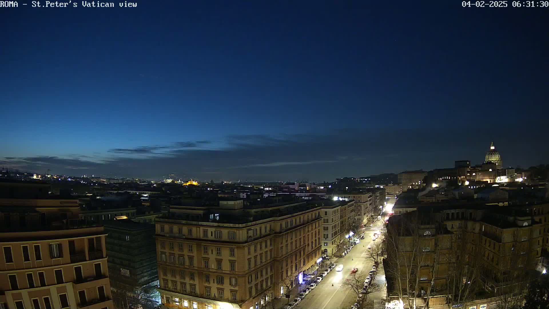 A nighttime, high-angle view of a city street with buildings illuminated, under a mostly clear, dark blue twilight sky.