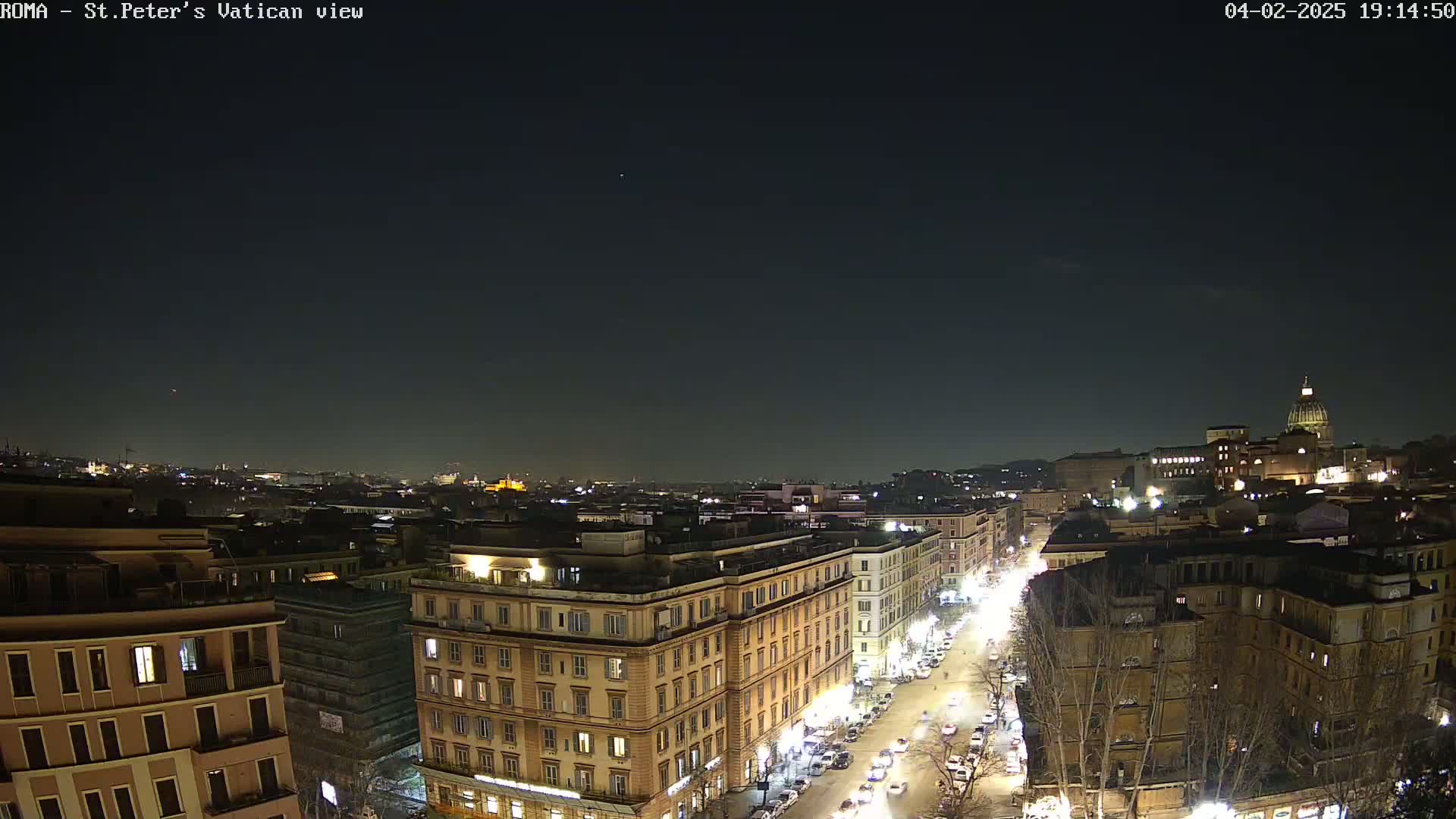 A nighttime view of a city street, lined with buildings, under a clear dark sky, with a distant dome-shaped structure visible in the background.