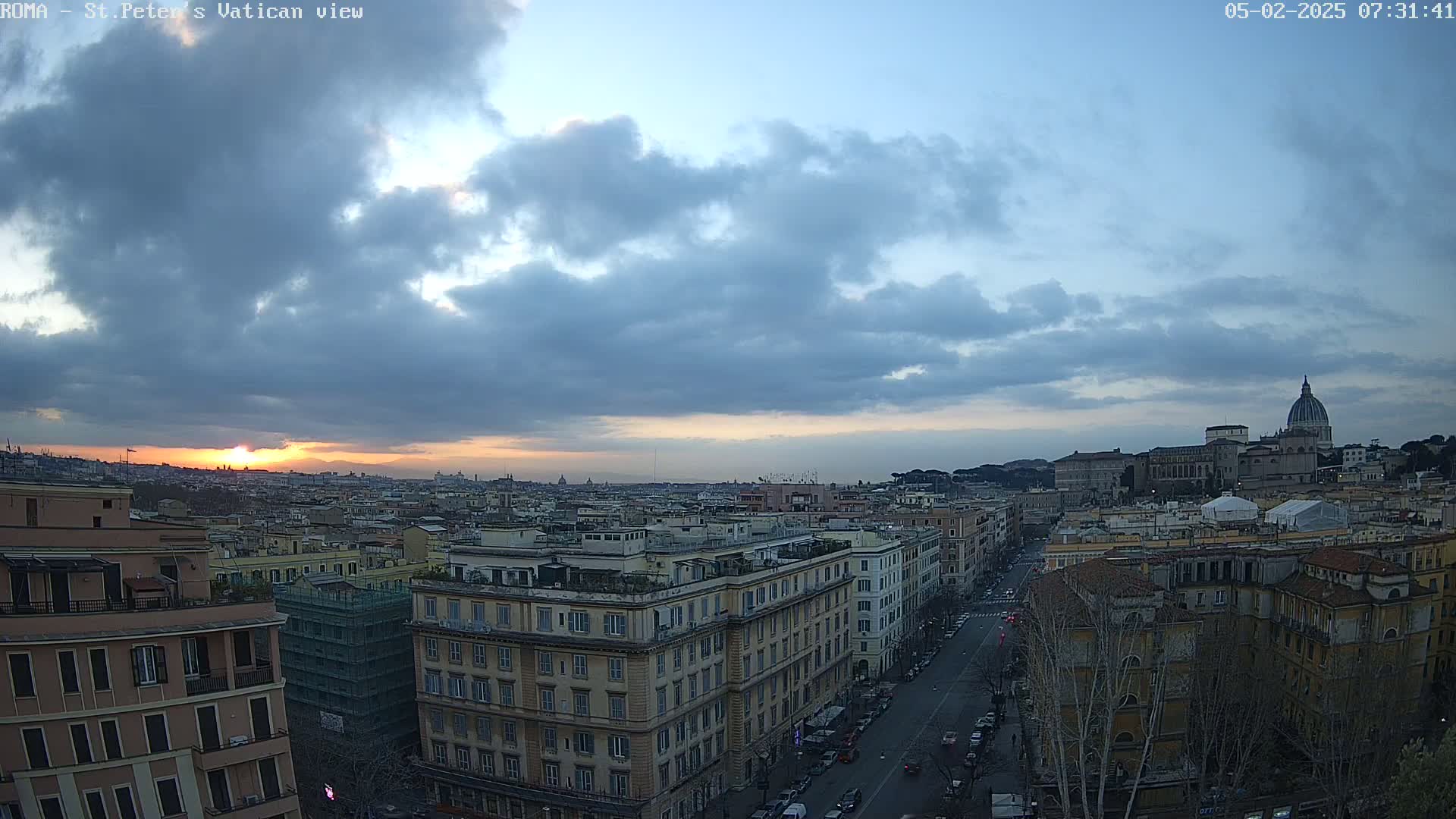 A sunset over a cityscape with cloudy skies, showing a wide expanse of buildings including St. Peter's Basilica in the distance.