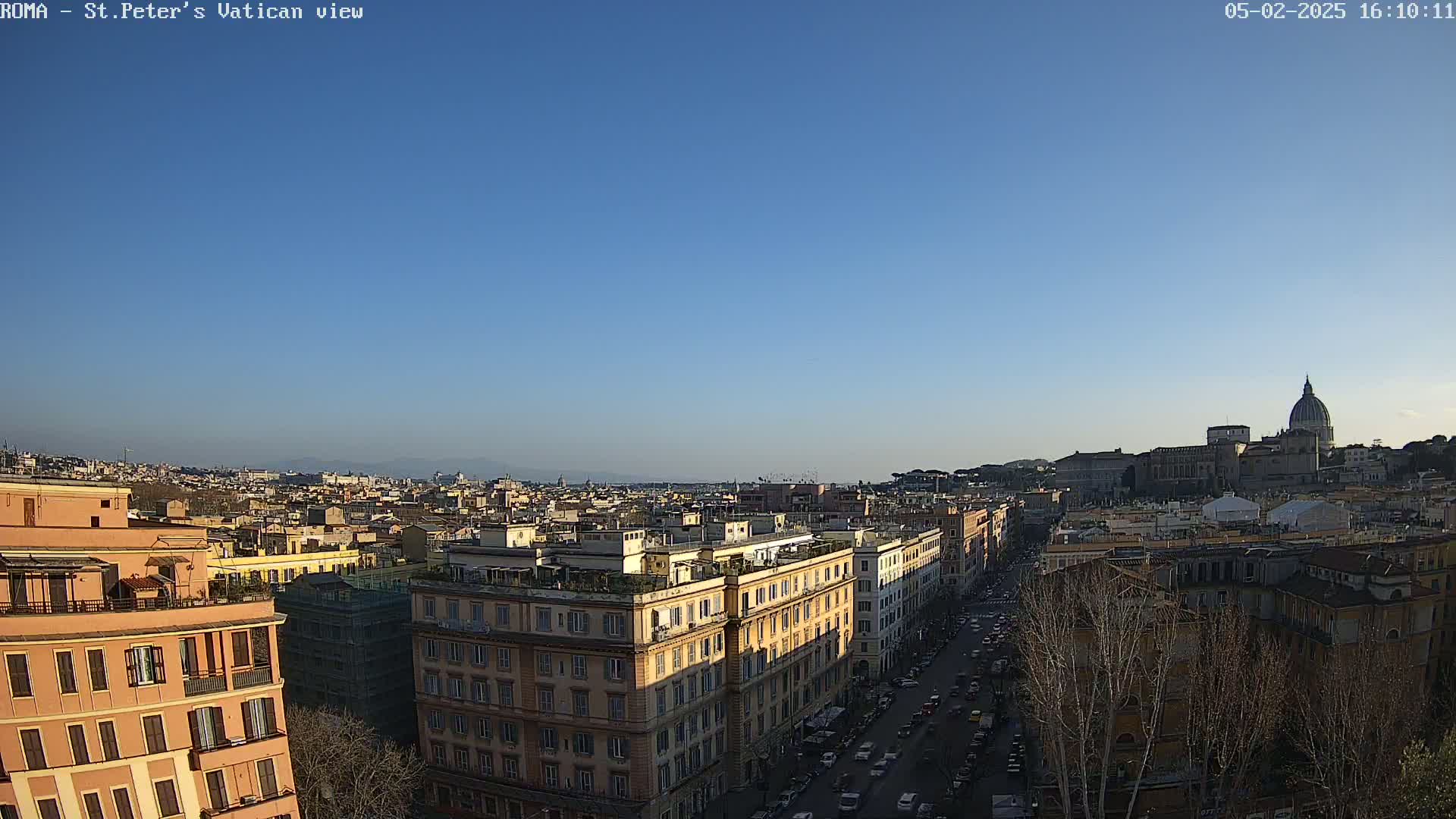 A high-angle, long shot on a clear, sunny day shows a cityscape with St. Peter's Basilica in the distance, and many buildings, streets, and trees in the foreground.