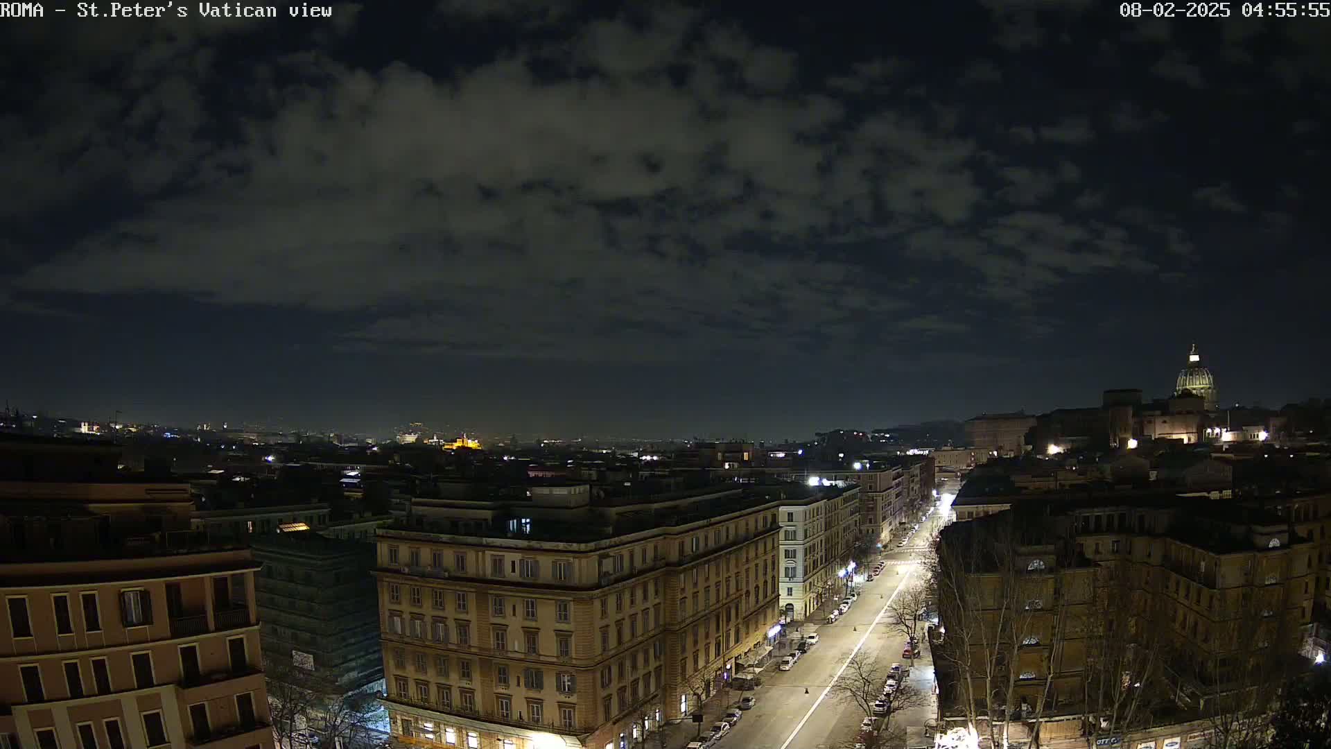 A nighttime view of a city street, with buildings illuminated and a dome-topped structure visible in the distance under a partly cloudy sky.