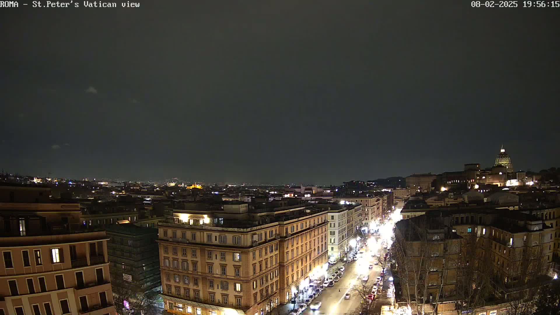 A nighttime, high-angle view of a city street lined with illuminated buildings, extending toward a distant, dimly lit cityscape under a dark, mostly clear sky.