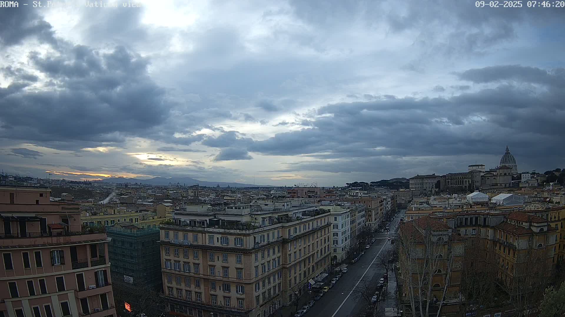 A cityscape under a cloudy sky, showing a wide expanse of buildings, a street, and a distant dome-topped structure, with mountains visible on the horizon.
