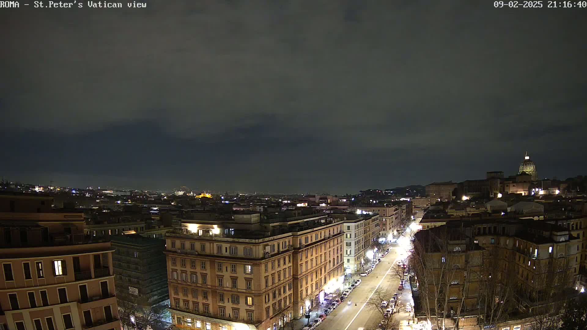 A nighttime view of a city, showing a street lined with buildings, with a distant dome-shaped structure visible under a dark, cloudy sky.