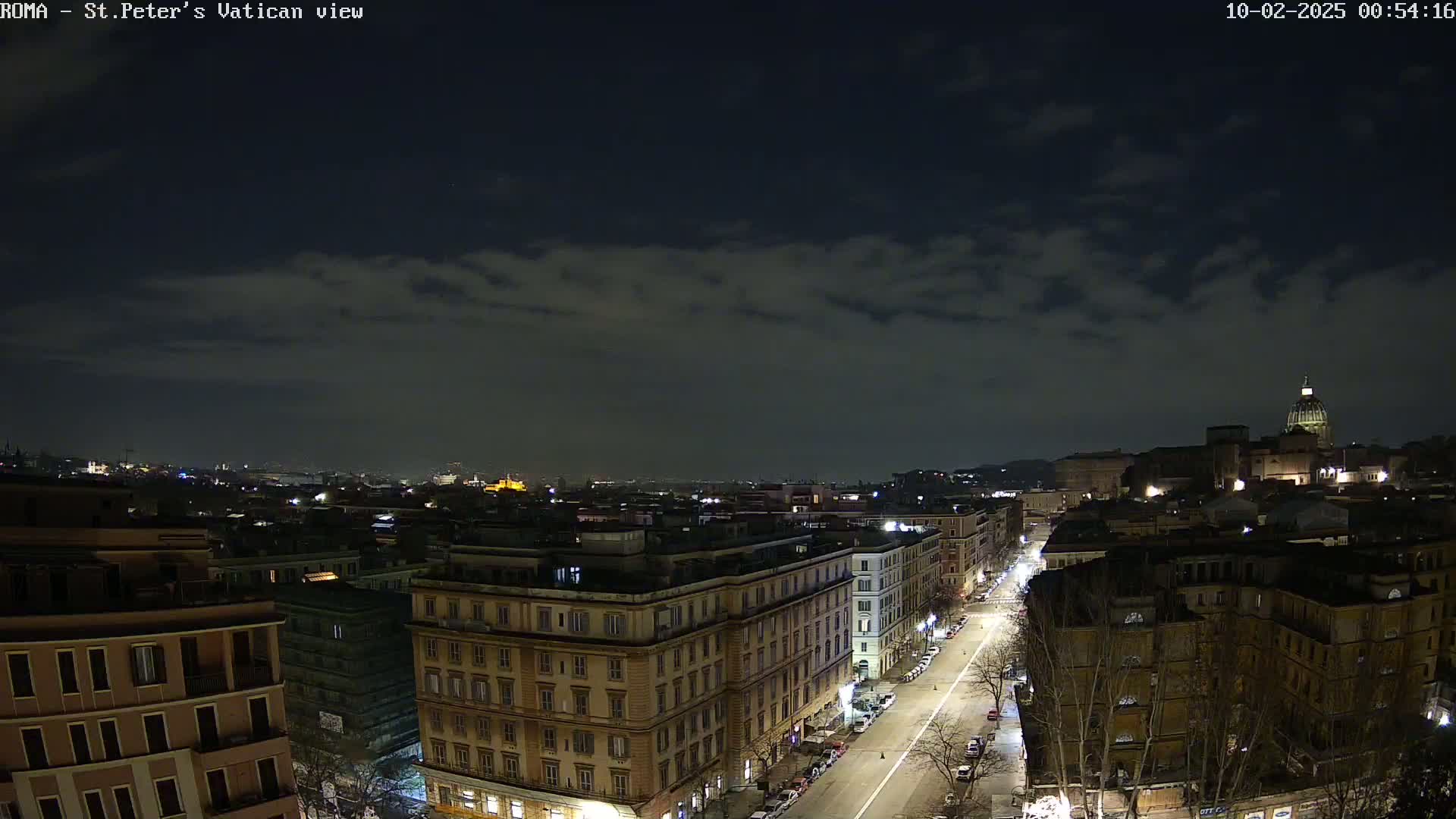 A nighttime view of a city street lined with buildings, extending towards a hilltop with a dome-shaped structure under a partly cloudy sky.
