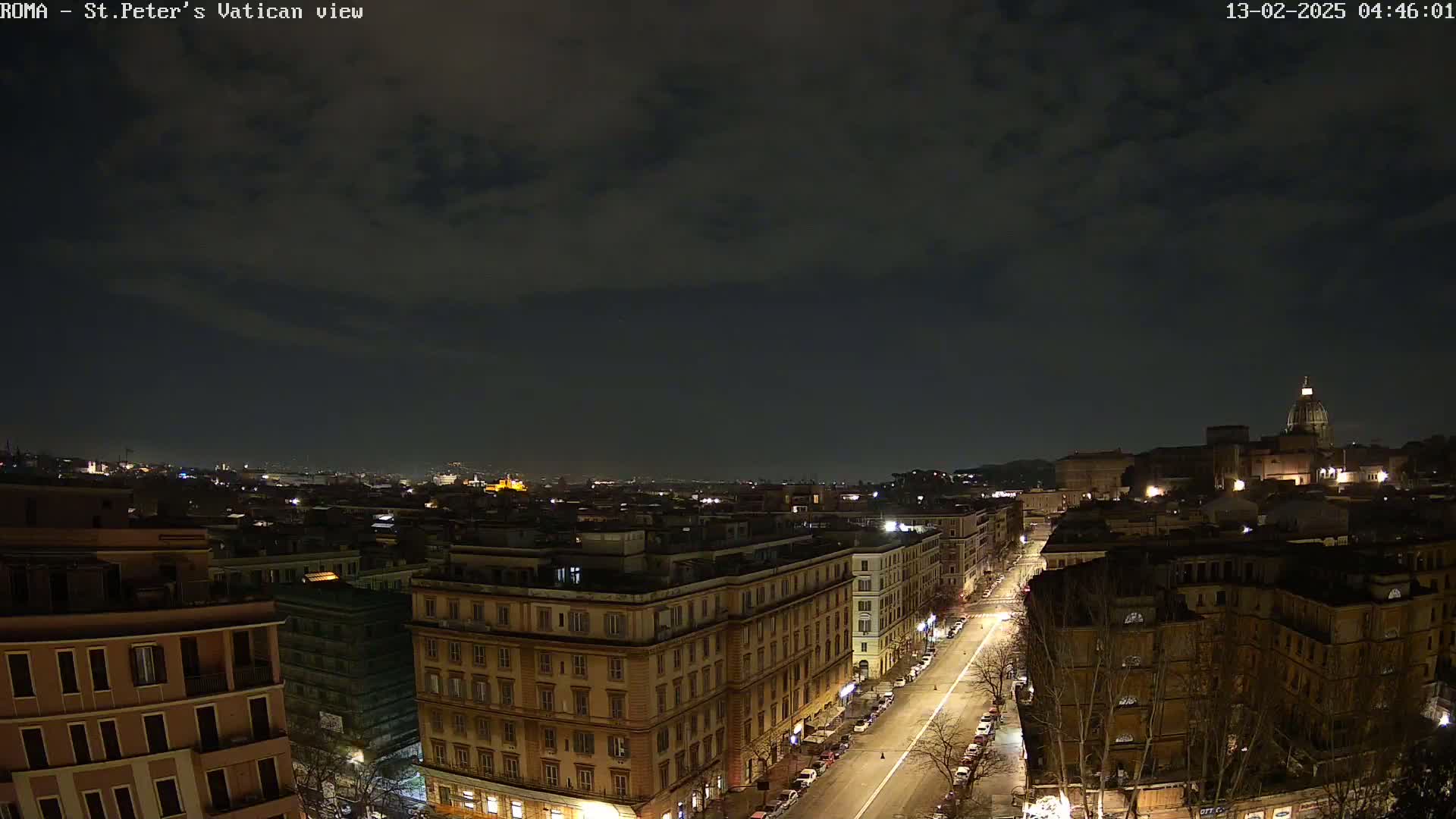 A nighttime, high-angle view of a city street lined with buildings, under a cloudy sky.