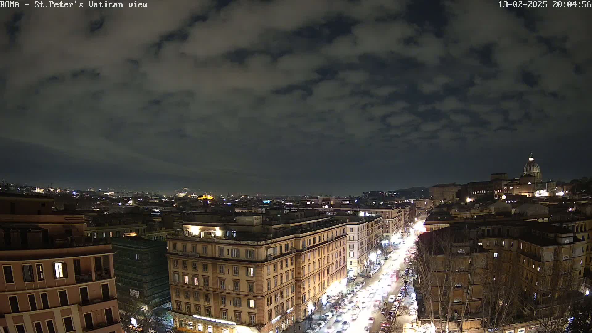 A nighttime cityscape with a partly cloudy sky shows a view of a city street with traffic and buildings, including a large structure with a dome in the distance.