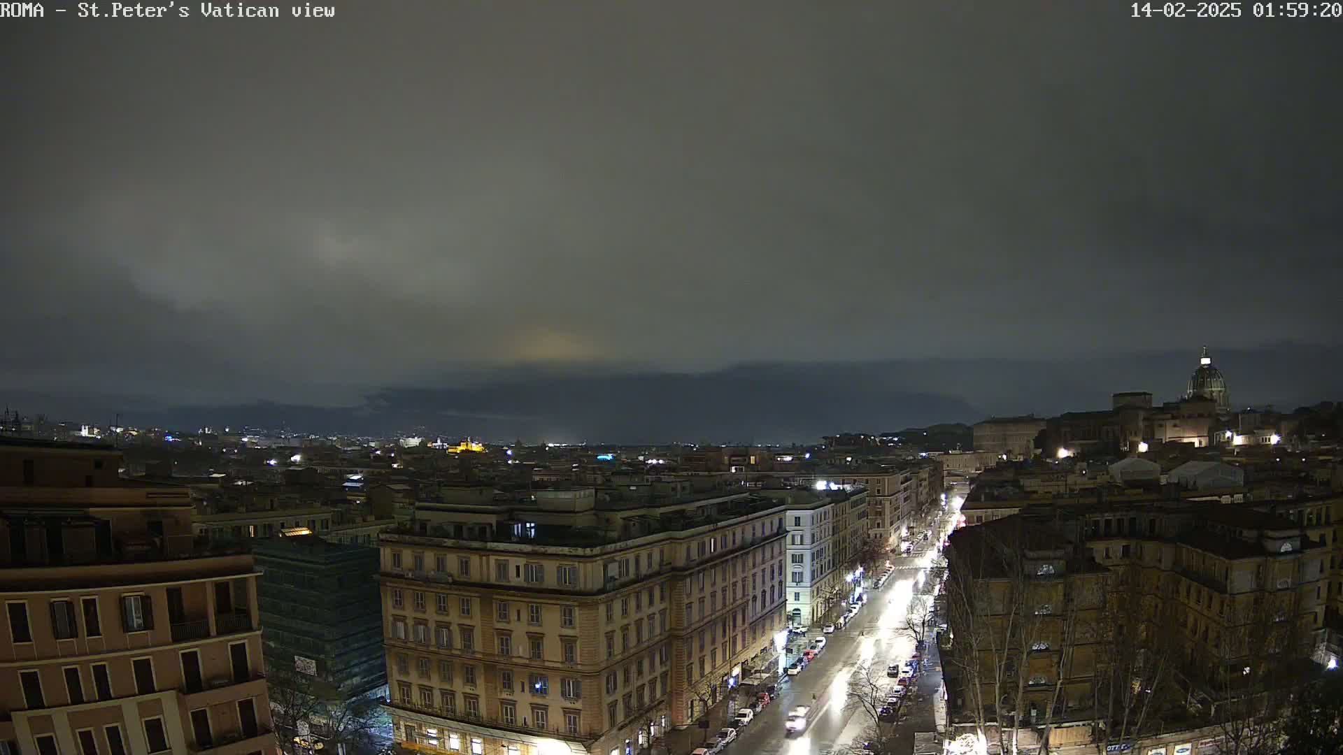 A nighttime, overcast view of a city, showing a street with cars and buildings lit up against a dark sky.