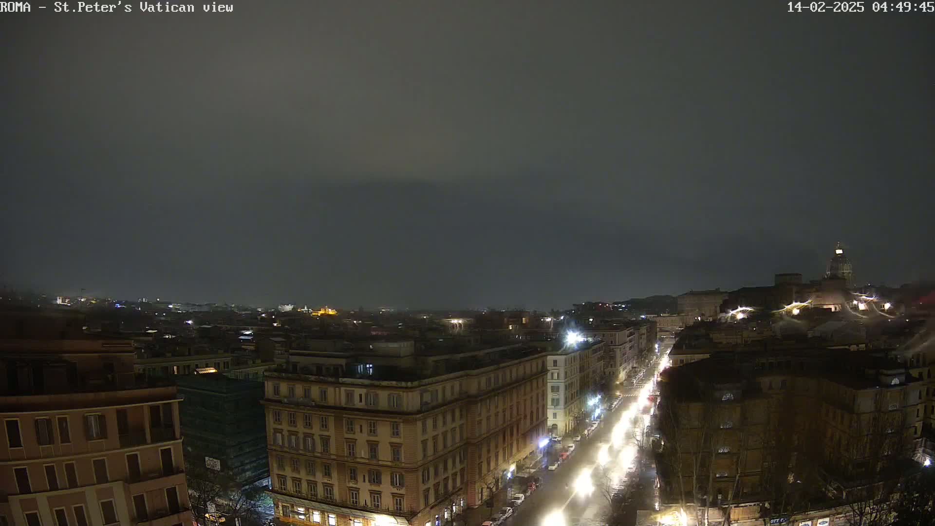A nighttime, overcast view of a city with illuminated streets and buildings, showing rain on the wet roads.