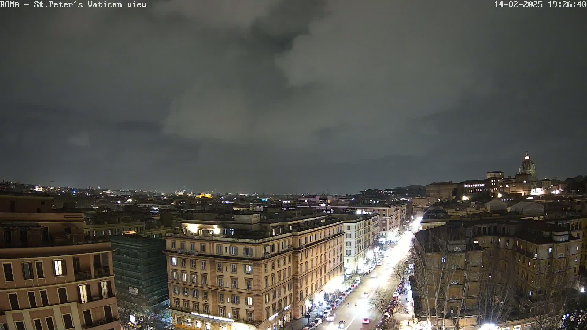A nighttime, overcast view of a city street scene, showing illuminated buildings and vehicles under a dark sky.