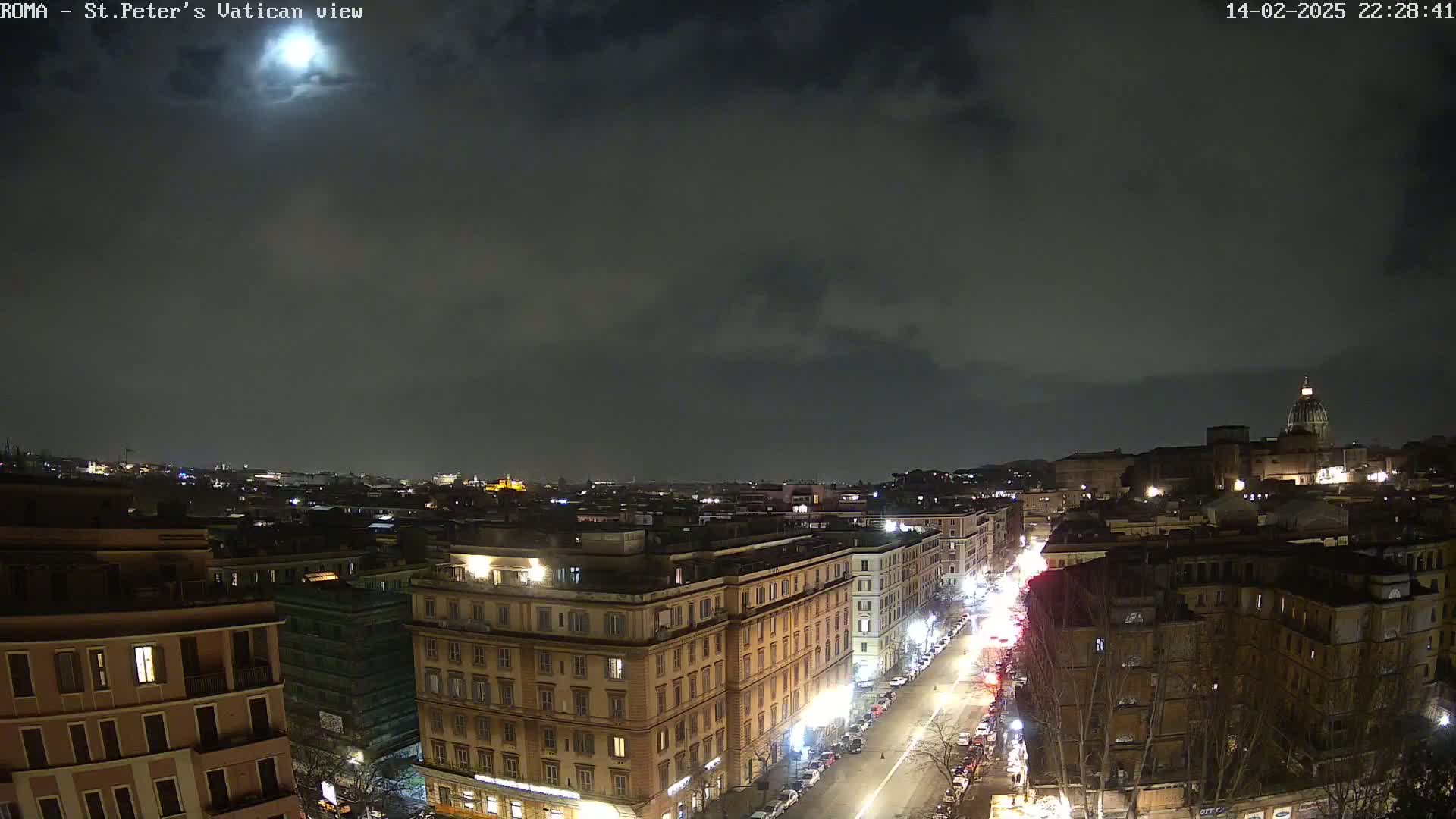 A nighttime, cloudy view of a city with a brightly lit street running through the center, showing buildings and distant lights under a mostly cloudy sky with a bright moon visible.