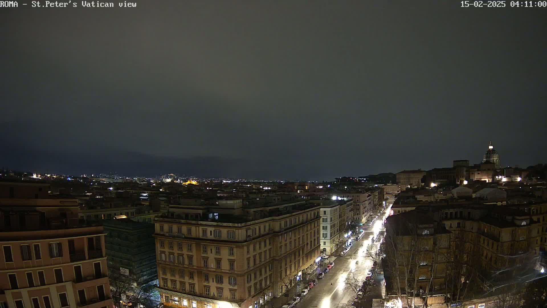A nighttime view of a city street illuminated by streetlights, with buildings lining both sides, extending to a cityscape in the distance under a dark, cloudy sky.