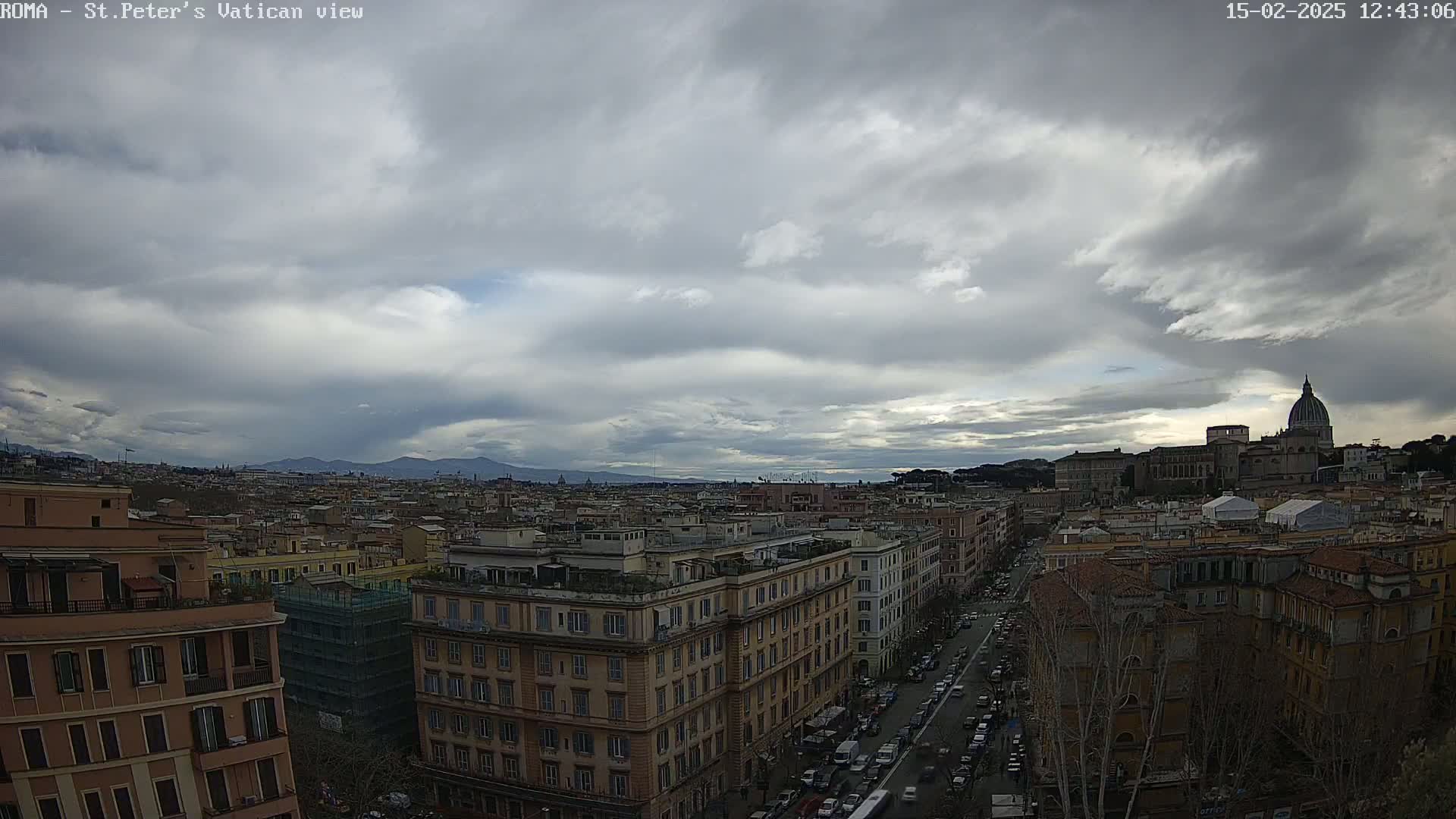 A view of Rome under a cloudy sky shows a cityscape with buildings and a distant dome, mountains visible in the background.