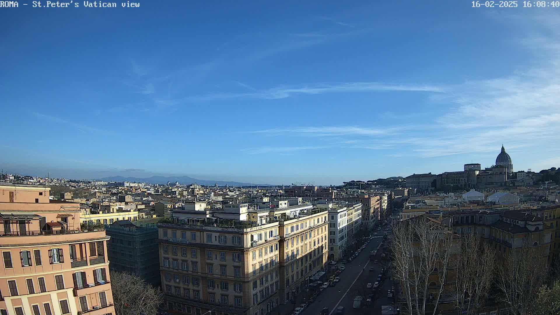 A cityscape view of Rome, under a clear blue sky, shows buildings, streets, and distant mountains.