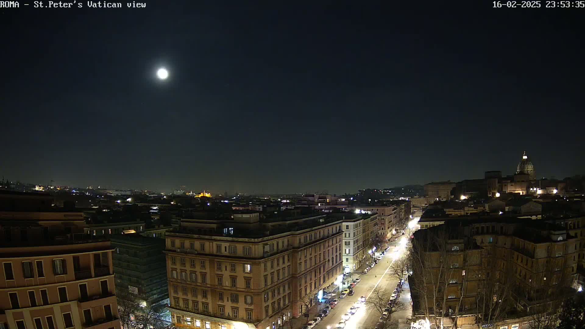 A nighttime aerial view of a city, illuminated by a bright moon, shows a street lined with buildings, with a large domed structure visible in the distance.