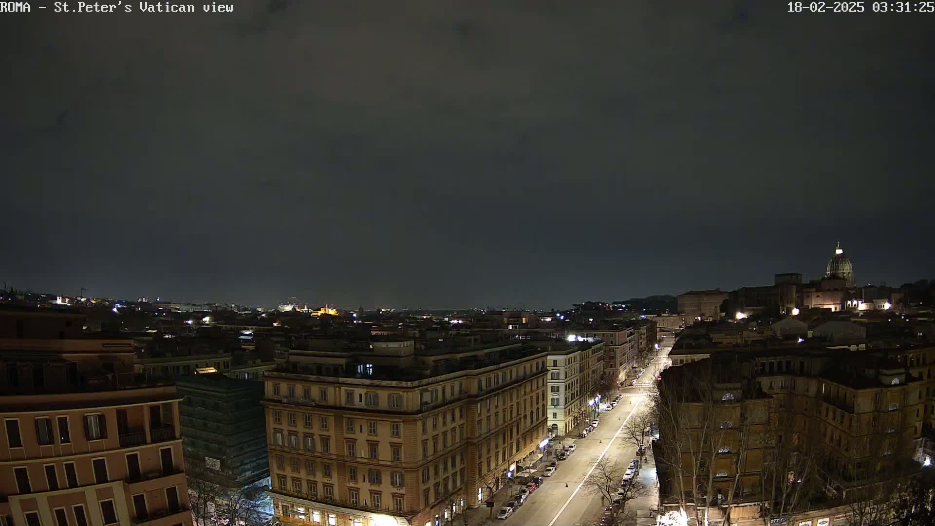 A nighttime, high-angle view of a city, showing a street with parked cars leading towards a large building with a dome under a dark, cloudy sky.