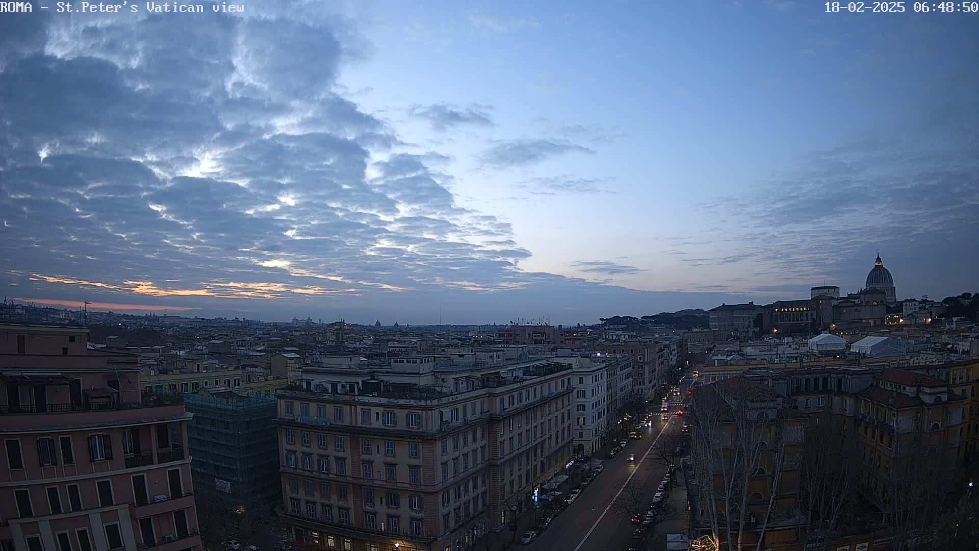 A cityscape at dawn under a mostly cloudy sky shows a street with cars, buildings, and in the distance, St. Peter's Basilica.