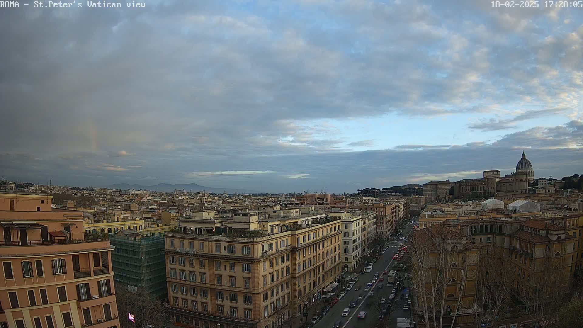 A partly cloudy sky with a faint rainbow hangs over a cityscape, showing a long street with traffic leading towards a large domed building in the distance.