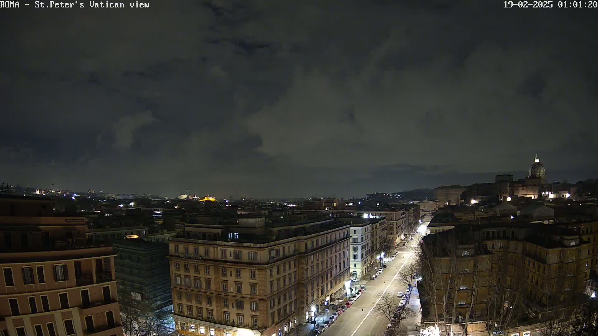 A nighttime, cloudy view of a city street lined with buildings, stretching toward a distant hilltop with illuminated structures under a dark sky.