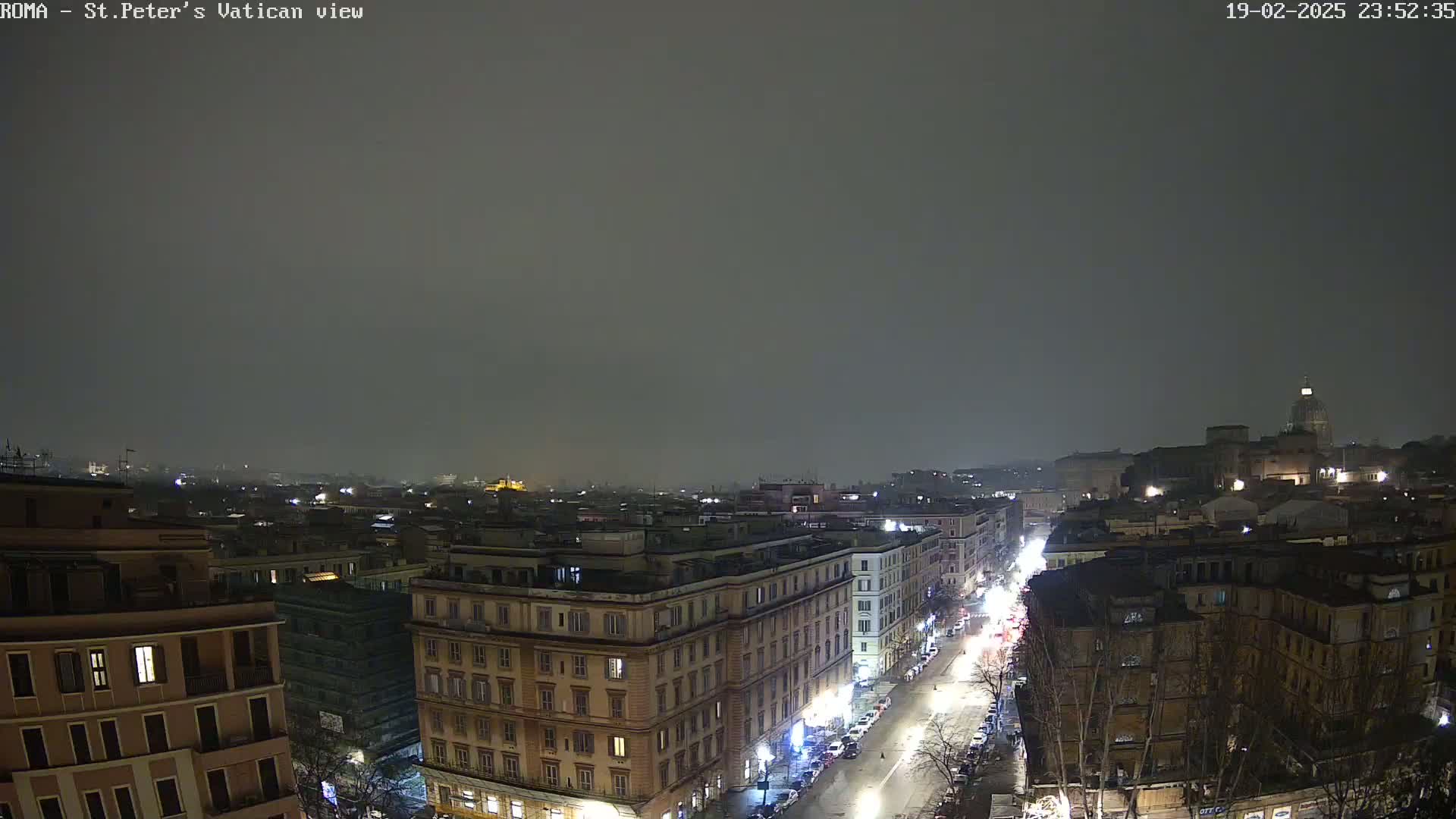 A nighttime, hazy view of a city street, lined with buildings, leading towards a distant structure with a dome under a dark sky.