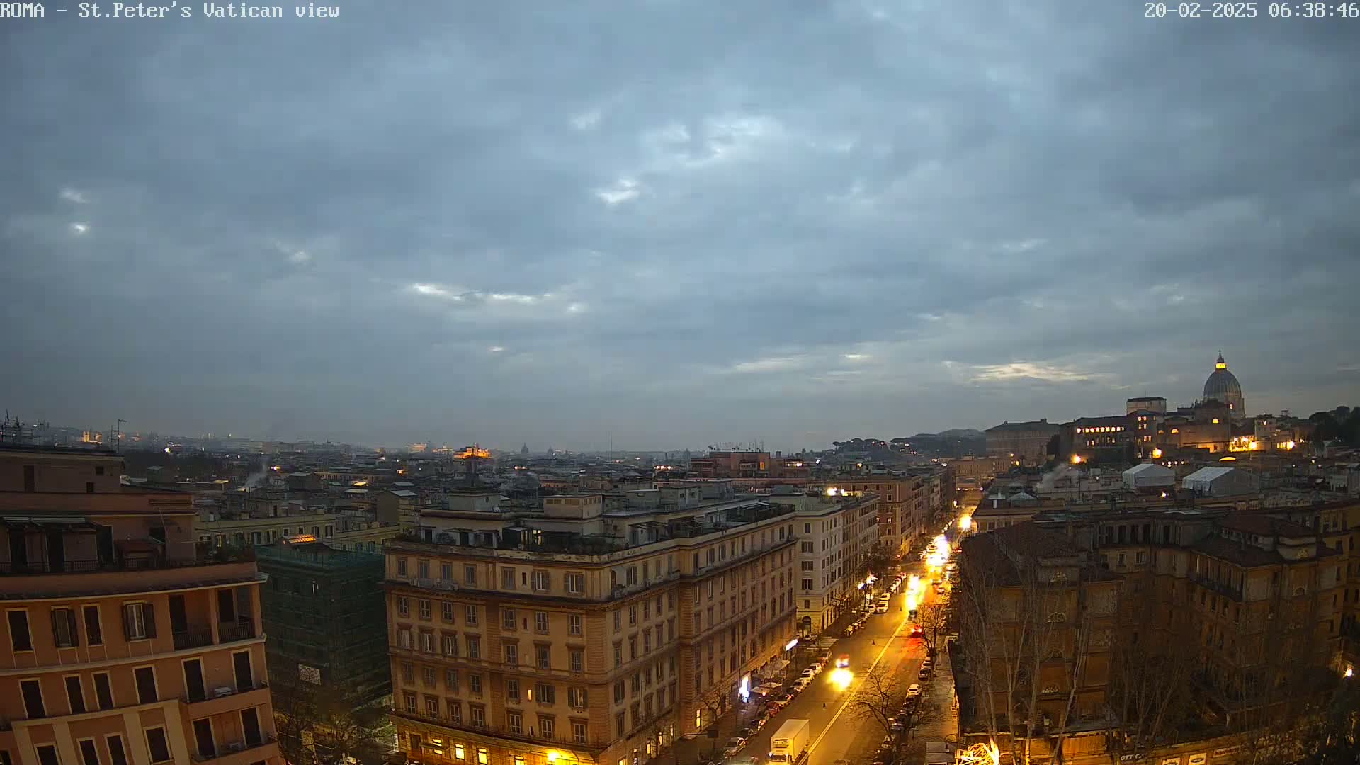 A nighttime view of a cityscape with a cloudy sky shows a street lined with buildings, illuminated by streetlights, and in the distance, the dome of St. Peter's Basilica.