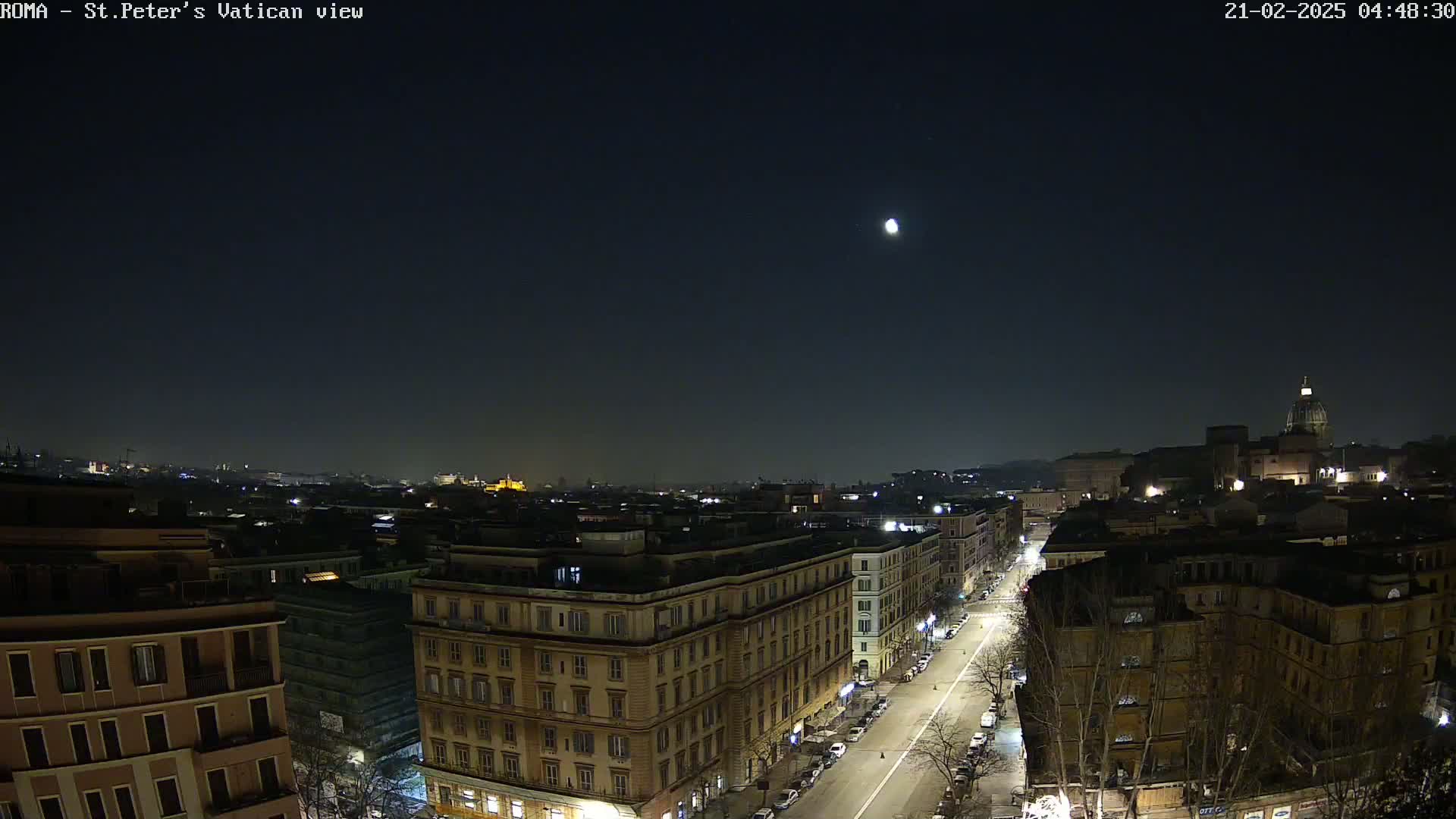 A nighttime aerial view of a city, possibly Rome, shows a moon in the sky above a street lined with buildings, with some illumination visible from streetlights and building windows.