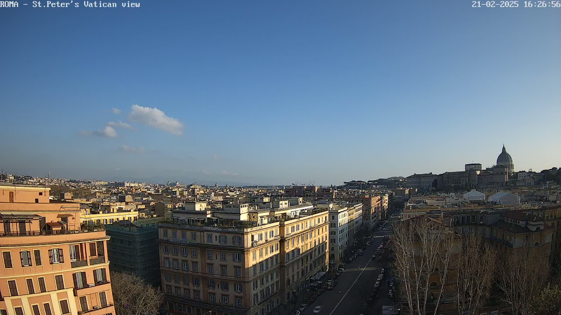 A clear, sunny day reveals a panoramic view of Rome, showcasing a cityscape with multi-story buildings, a wide street, and St. Peter's Basilica in the distance.