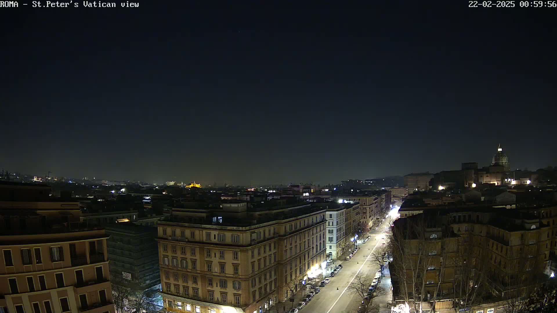 A nighttime, high-angle view of a city street scene, showing buildings and a road with parked cars, under a clear, dark sky.