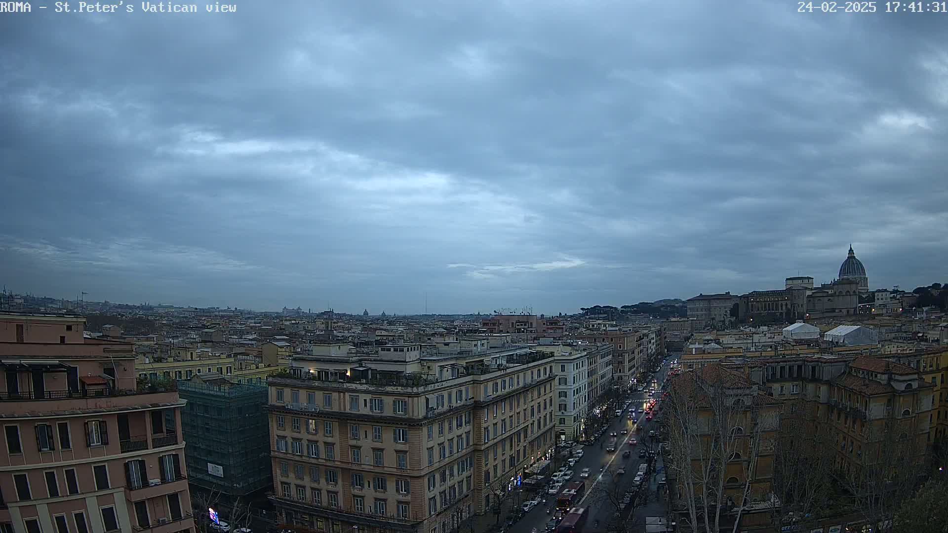 A view of a city, under a cloudy sky, shows St. Peter's Basilica in the distance.