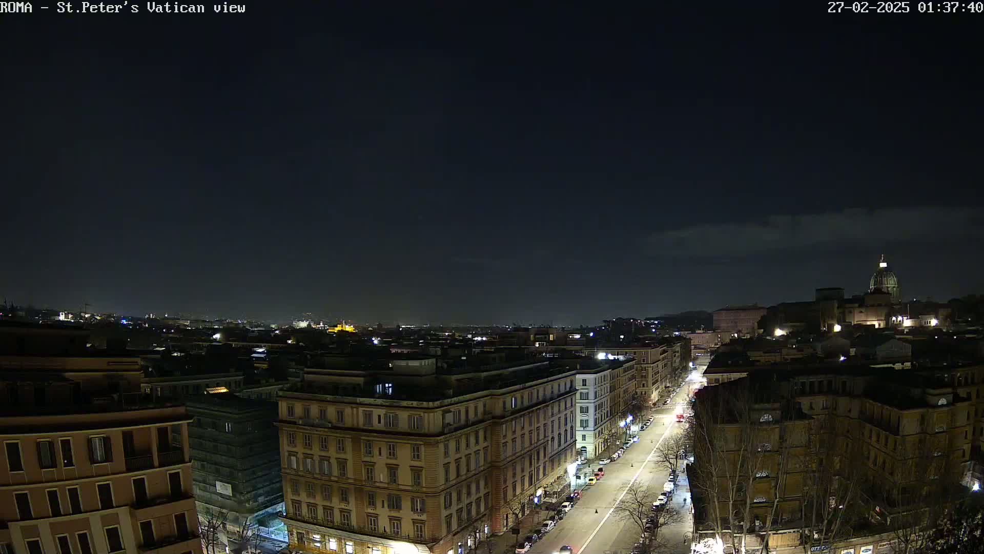 A nighttime, high-angle view of a city street, lined with buildings, leading towards a large dome-topped structure in the distance under a mostly clear sky.