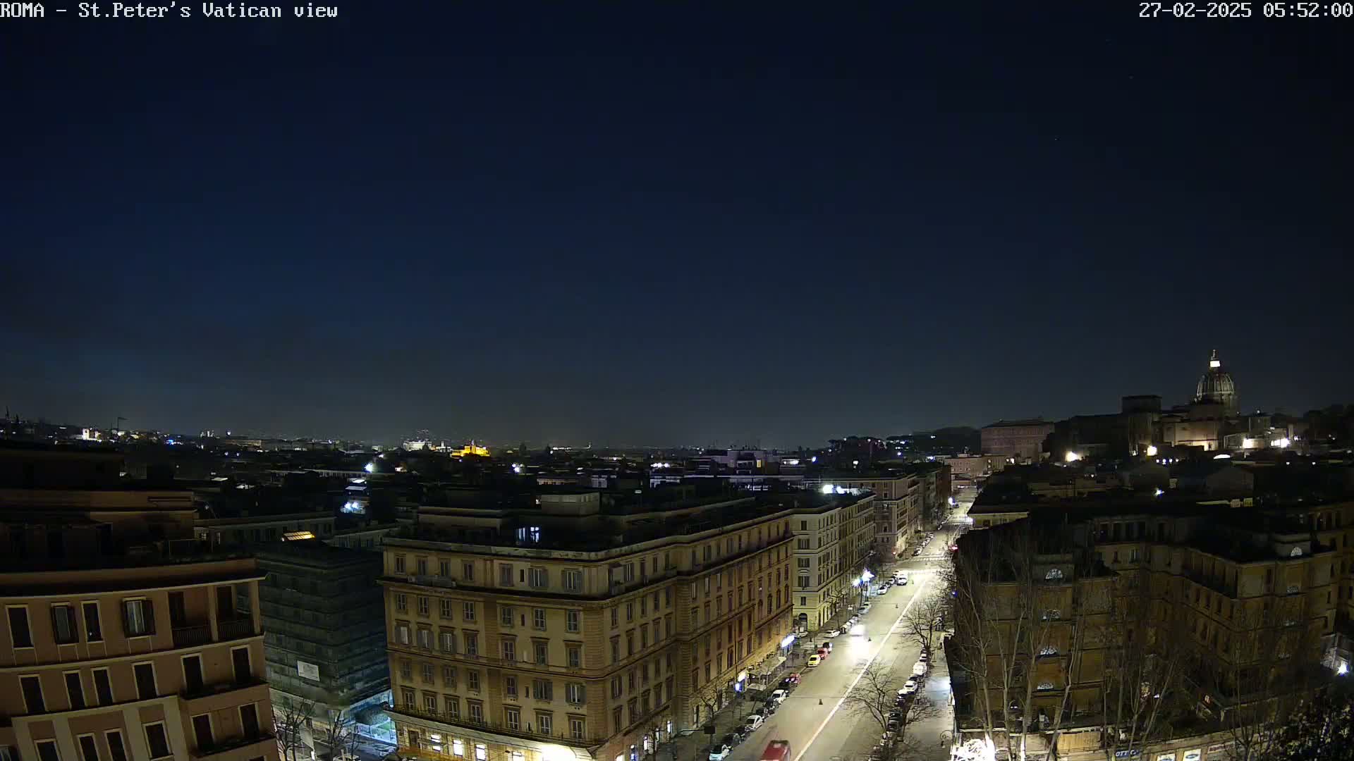 A nighttime, high-angle view of a city, showing illuminated streets and buildings, with a dome-shaped structure visible in the distance under a clear, dark sky.