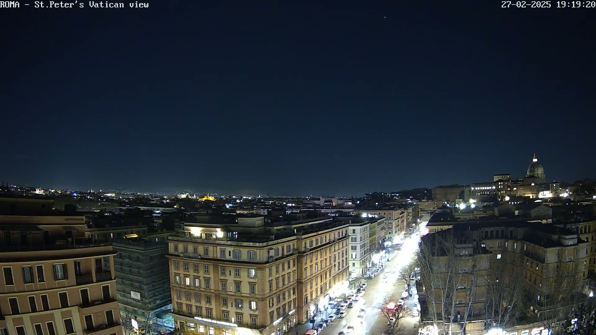 A nighttime, aerial view of a city, showing a street with cars and buildings illuminated under a clear, dark sky.