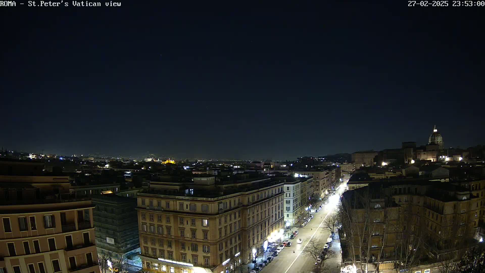 A nighttime, high-angle view of a city street, with buildings illuminated, leading towards a distant, larger structure topped with a dome under a clear, dark sky.