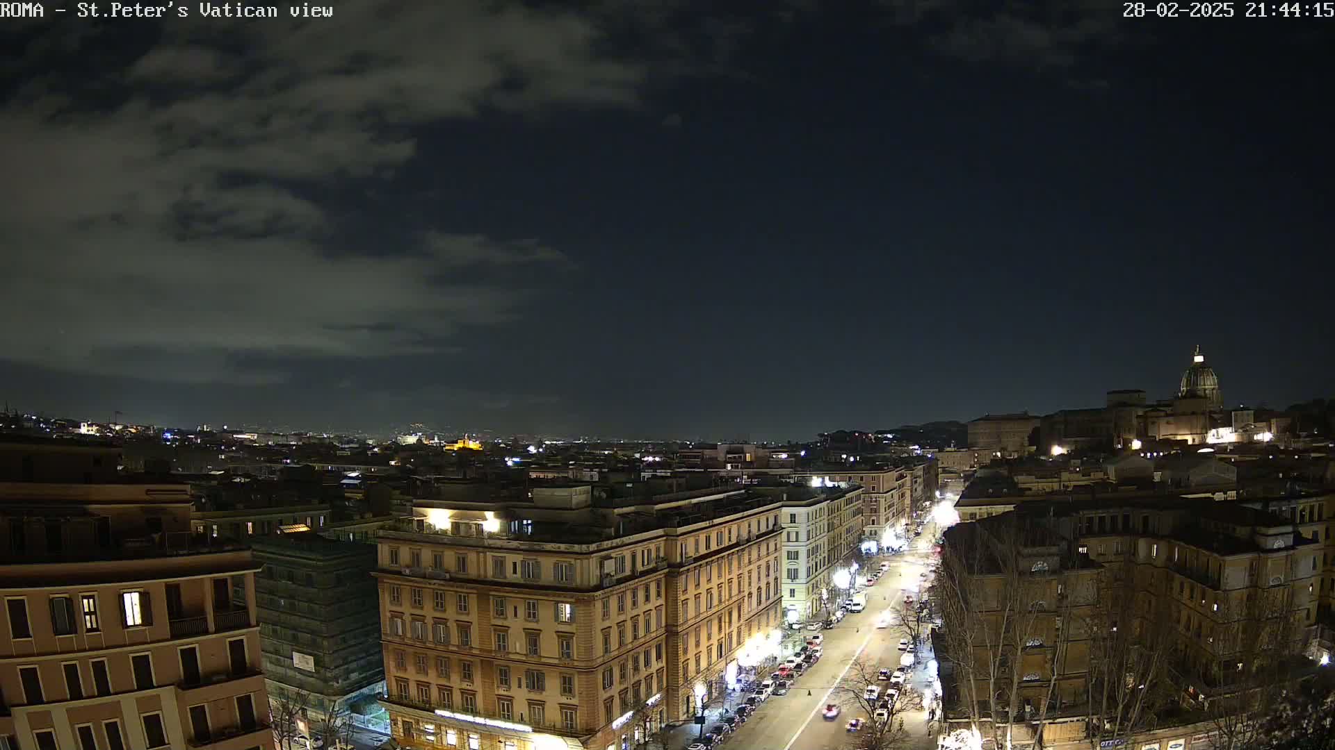 A nighttime view of a city, under a partly cloudy sky, shows a street with cars, buildings lit up, and in the distance, a large domed structure.