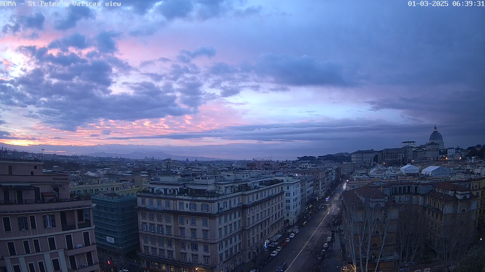 A cityscape at dawn under a mostly cloudy sky with pink and purple hues near the horizon.