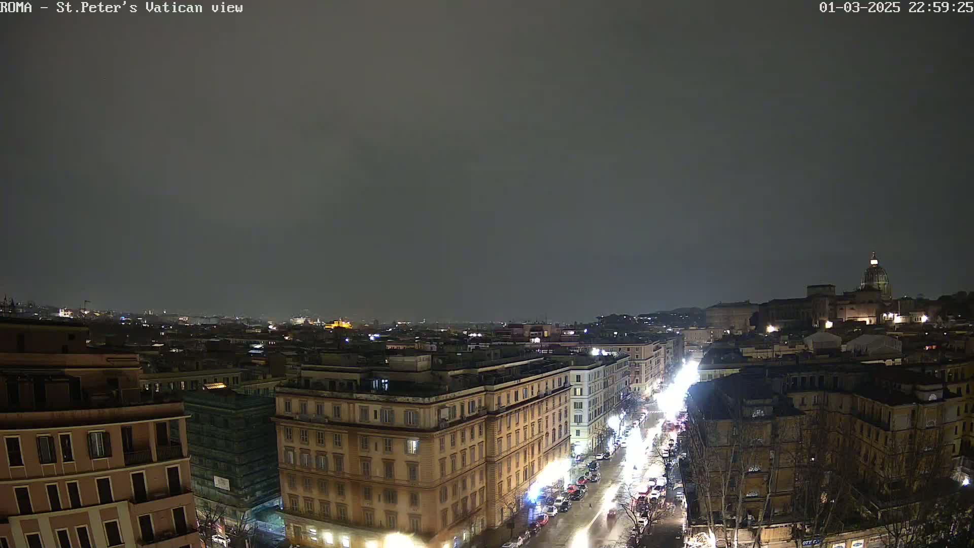 A nighttime view of a city street with buildings lit up under a dark, overcast sky.