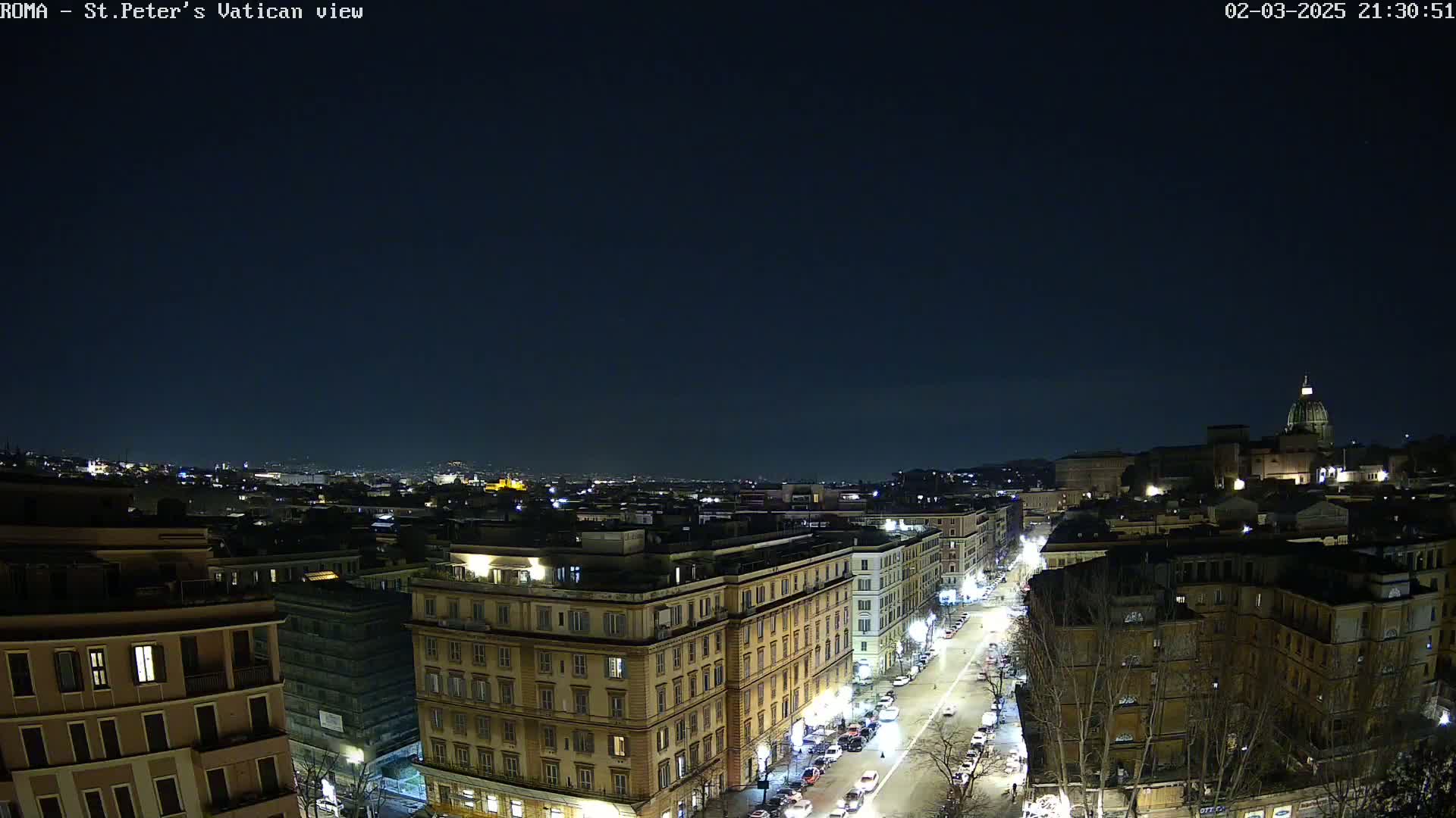 A nighttime, high-angle view of a city, showing a street with cars and illuminated buildings extending toward a distant dome-topped structure under a clear, dark sky.