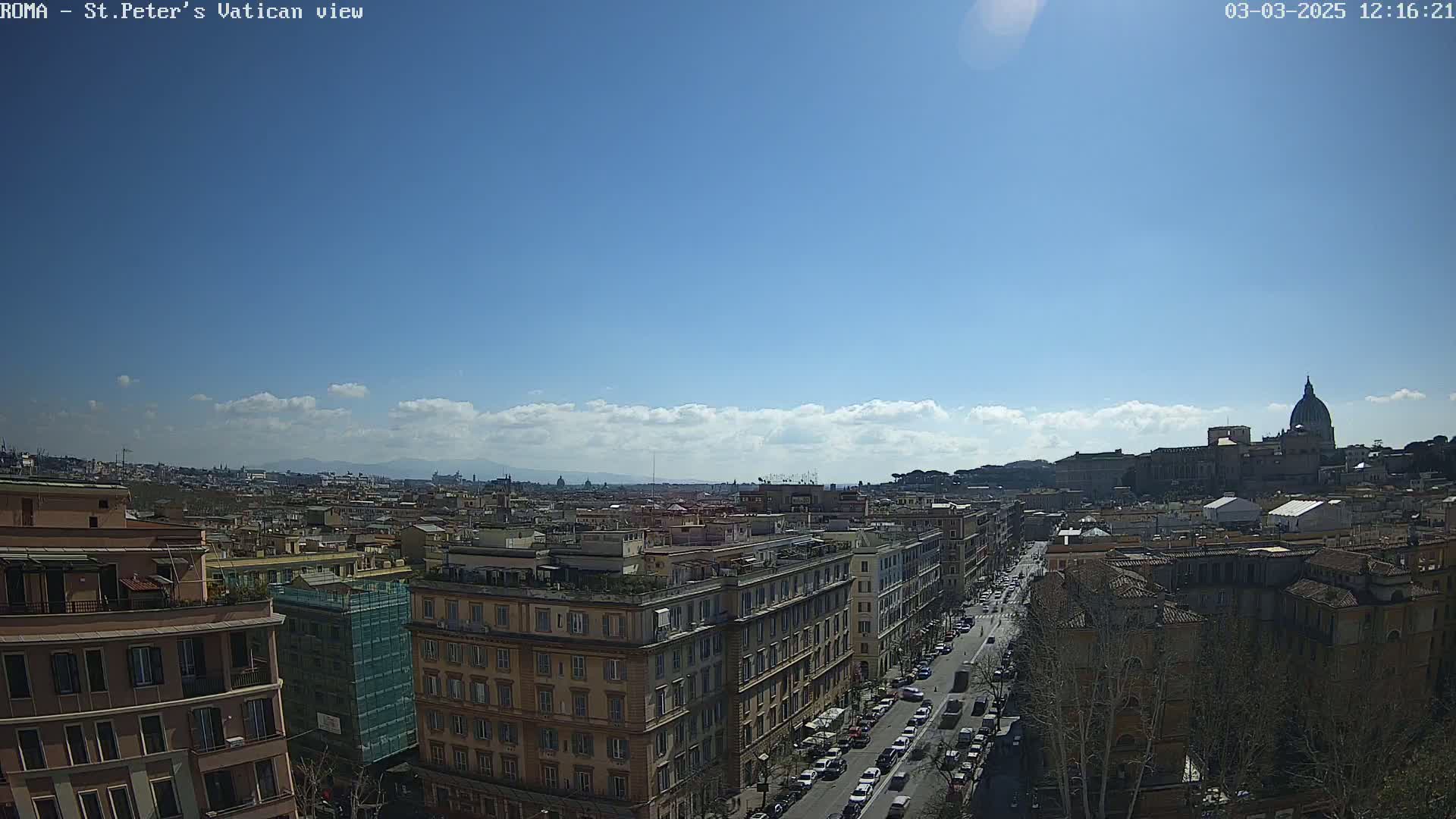 A sunny day reveals a panoramic view of Rome, featuring a cityscape of buildings and a street with cars, with the dome of St. Peter's Basilica visible in the distance.
