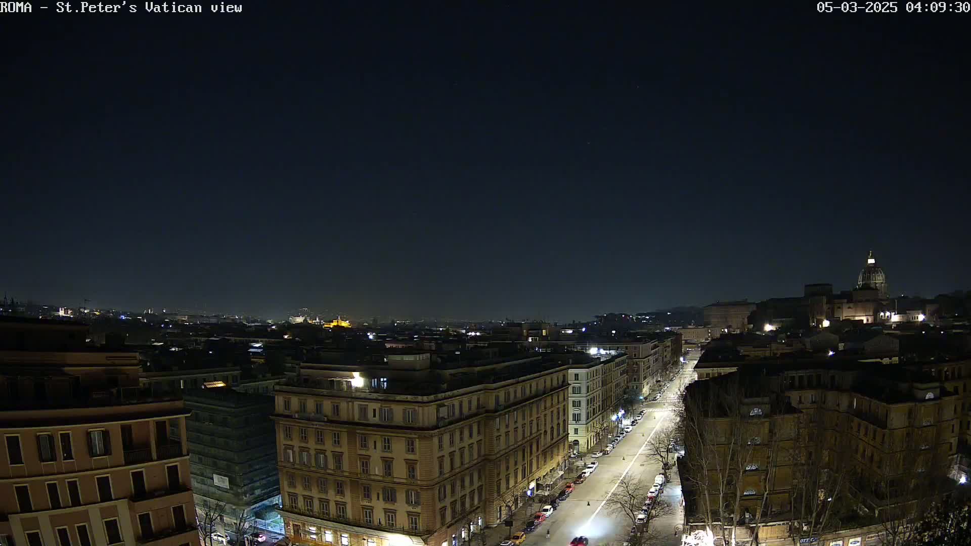 A nighttime, high-angle view of a city street lined with buildings, with a larger structure, possibly a church, visible in the distance under a clear, dark sky.