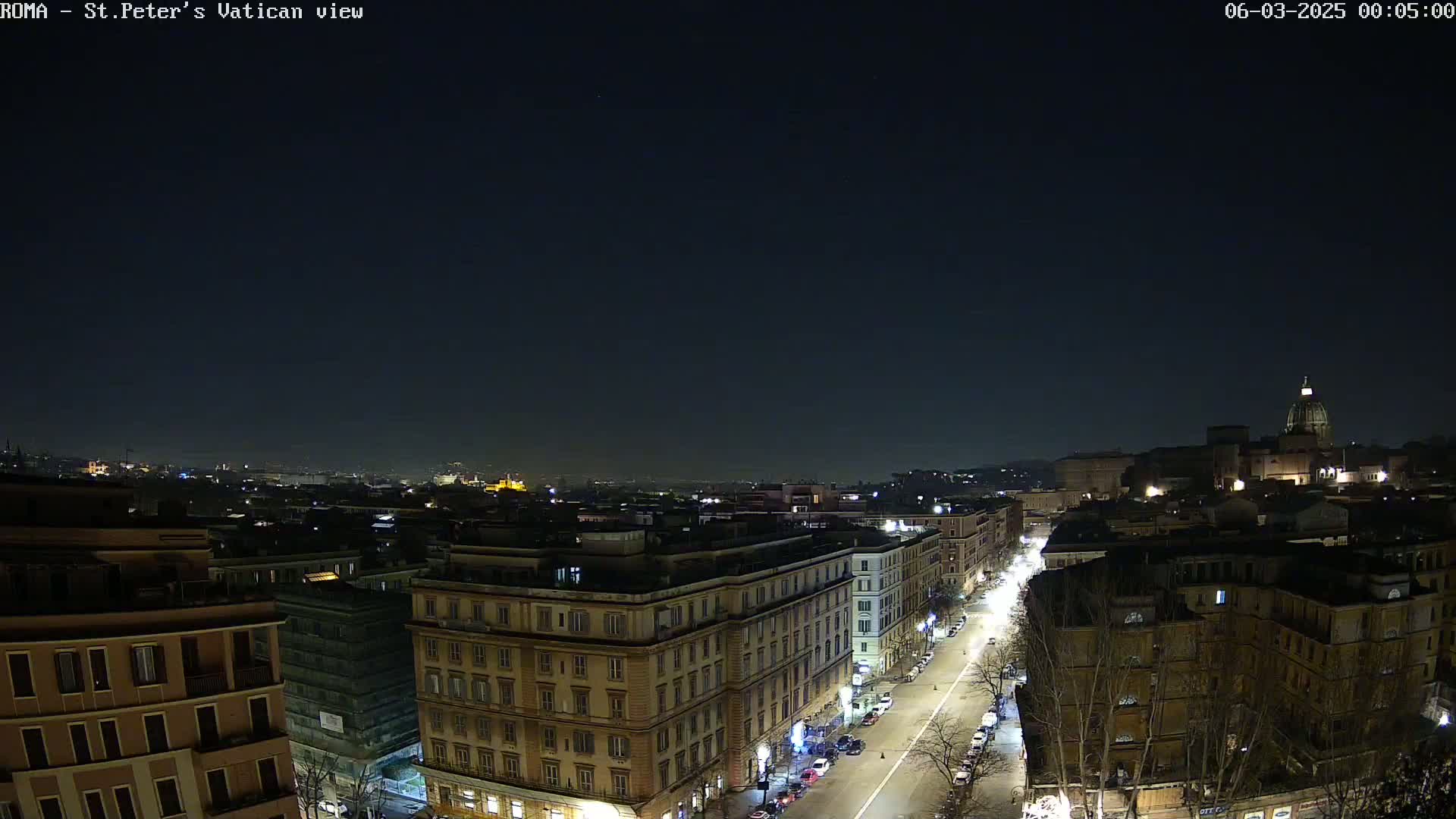 A nighttime aerial view of a city, showing a street lined with buildings under a clear, dark sky.