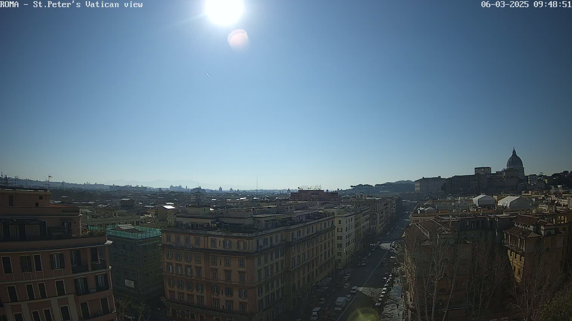 A sunny, clear day reveals a panoramic view of Rome, with the dome of St. Peter's Basilica visible in the distance, overlooking a cityscape of buildings and streets.